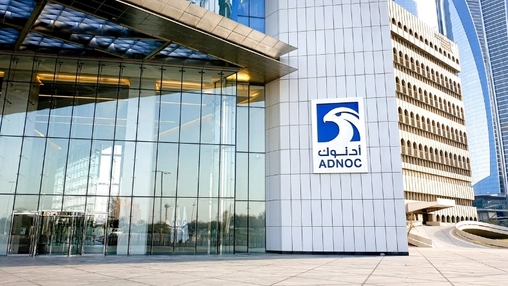 ADNOC headquarters with glass facade and company logo on a white tiled pillar, other towers visible behind