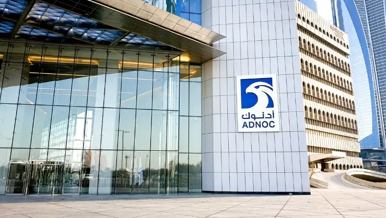 ADNOC headquarters with glass facade and company logo on a white tiled pillar, other towers visible behind